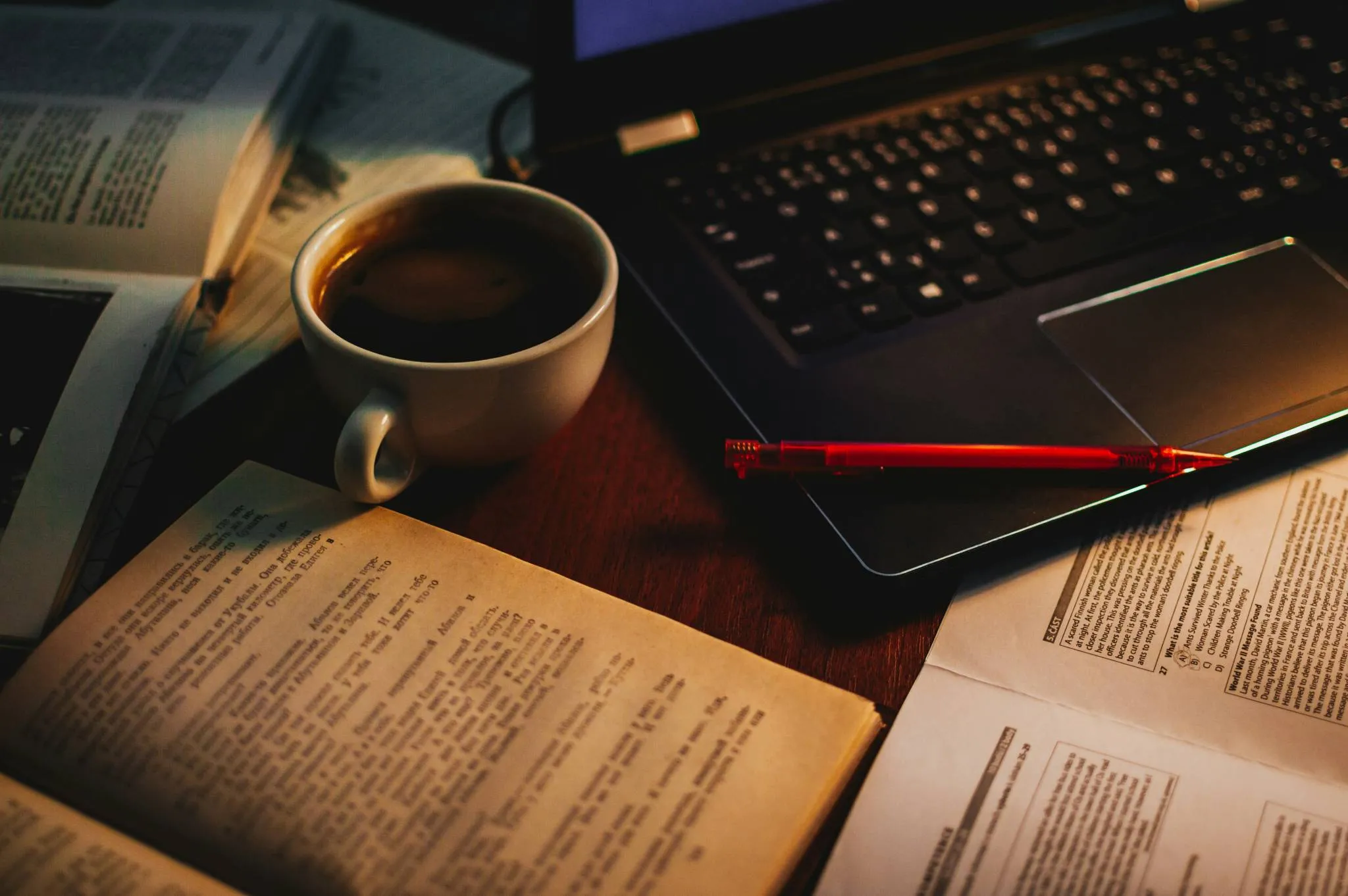 Open books, a cup of coffee, a red pen and a laptop on a wooden desk